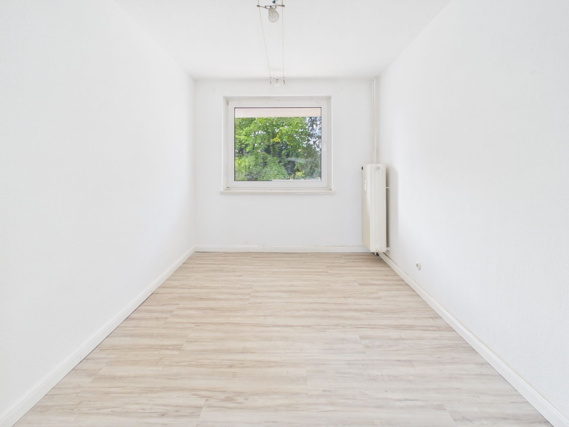 Heller, leerer Raum mit weißem Wänden, großem Fenster und hellem Holzboden.