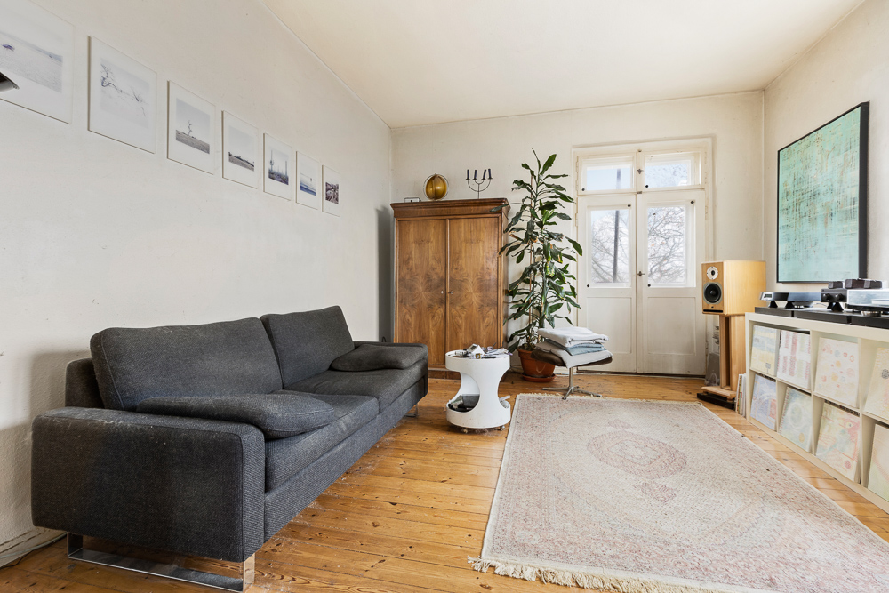 Wohnzimmer mit grauem Sofa, großem Teppich, Holzschrank, Pflanze und Bildern an der Wand.