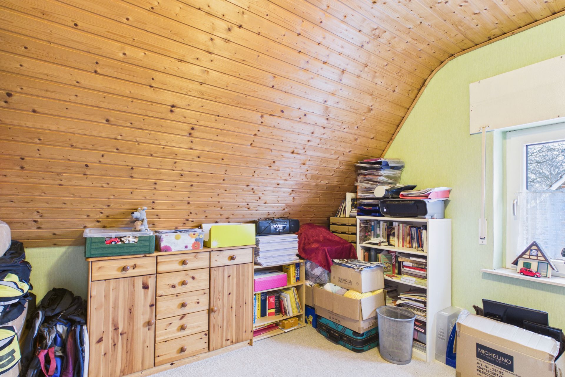 Zimmer mit Holzdecke, Regal, Kommode, Kartons und Büchern neben einem Fenster.