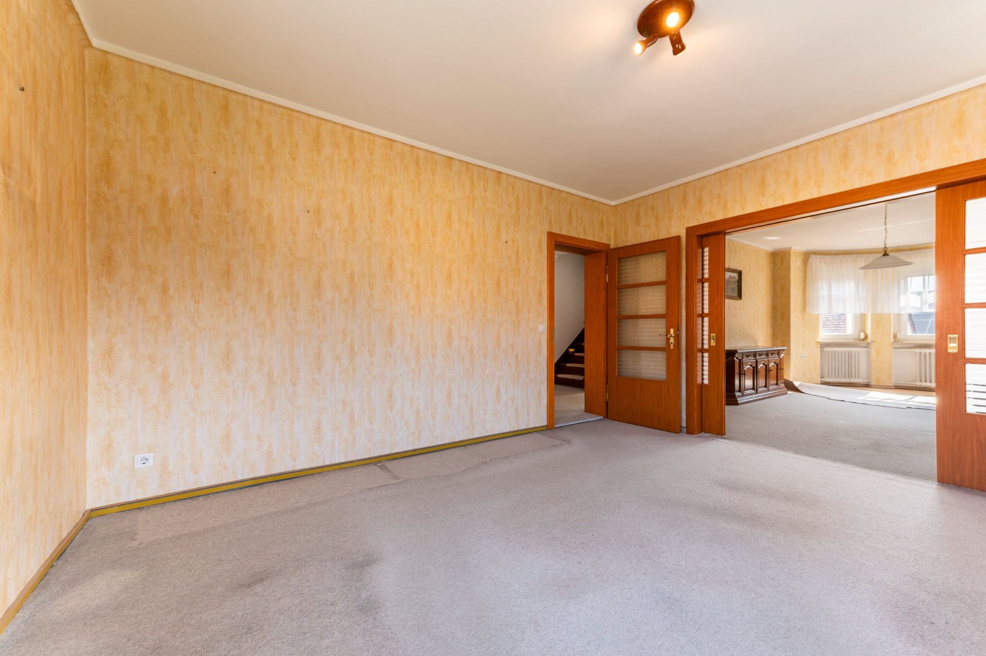 Leeres Wohnzimmer mit Teppichboden, gelben Wänden und Holztüren, Übergang zu einem weiteren Raum.