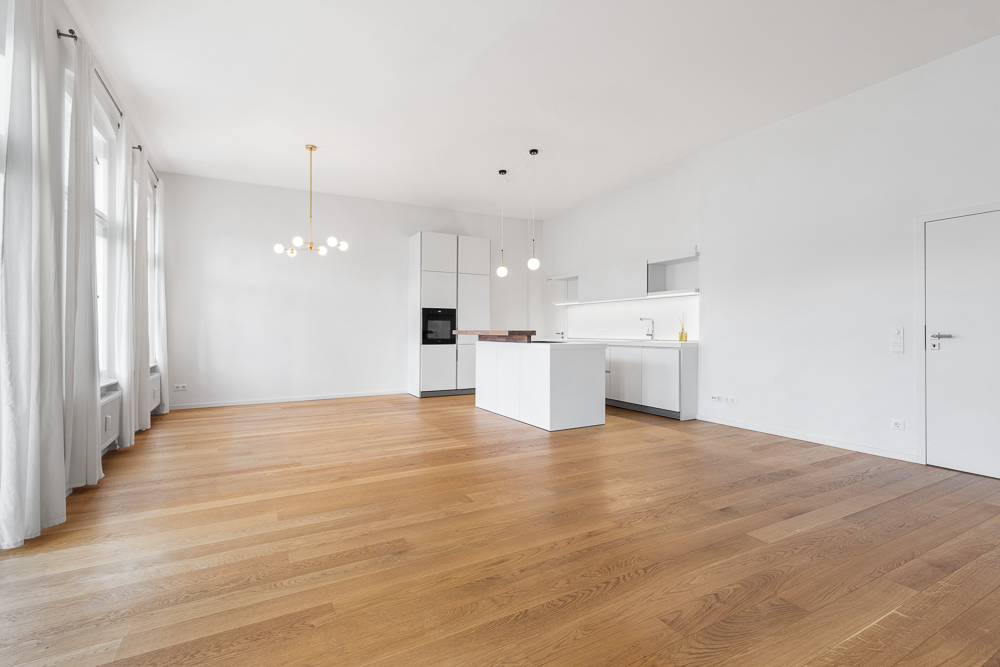 Helles, modernes Wohnzimmer mit offener Küche, Holzboden und großen Fenstern mit weißen Vorhängen.