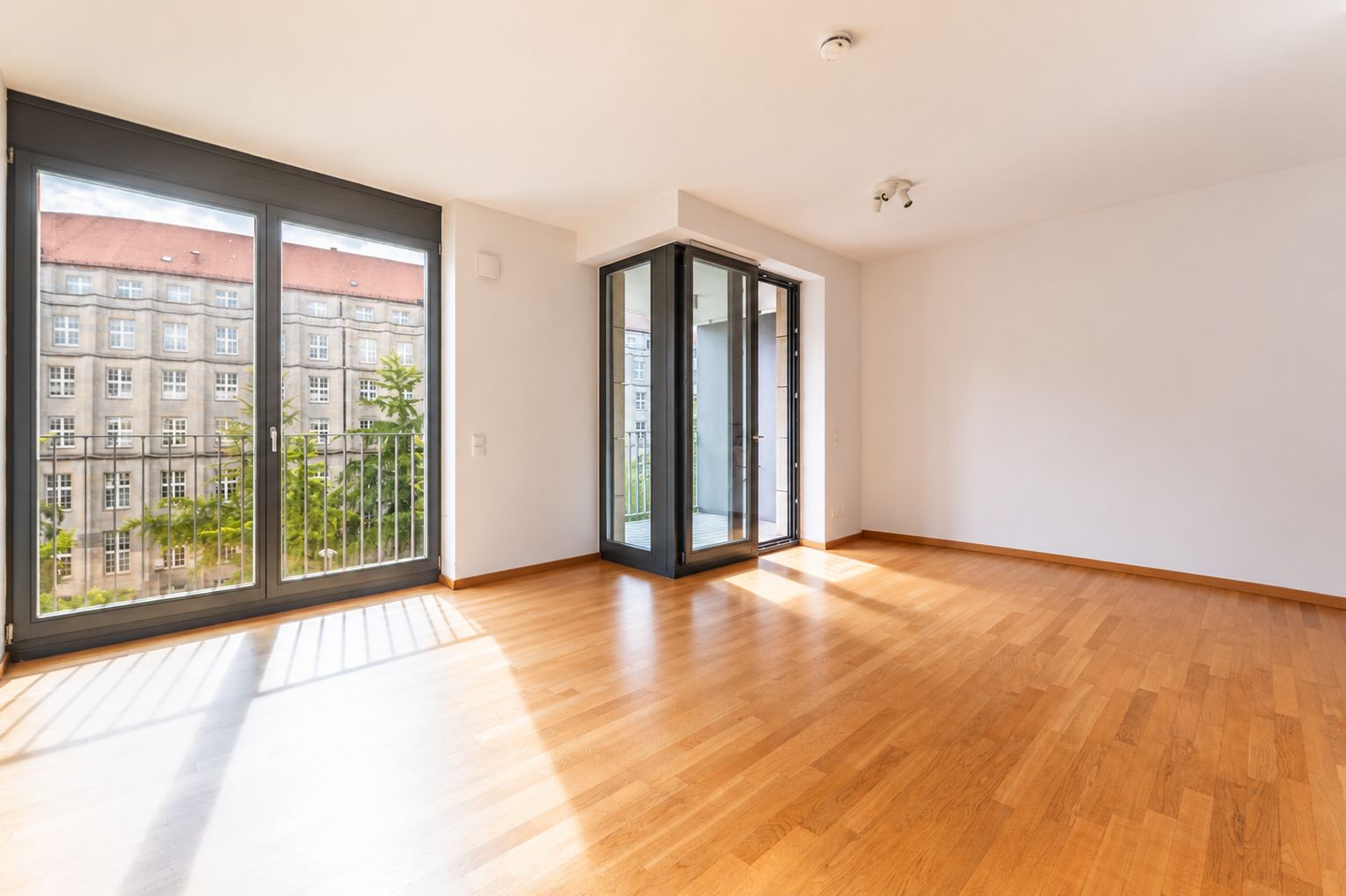 Helles Zimmer mit großem Fenster, Holzboden und Zugang zu einem verglasten Balkon.