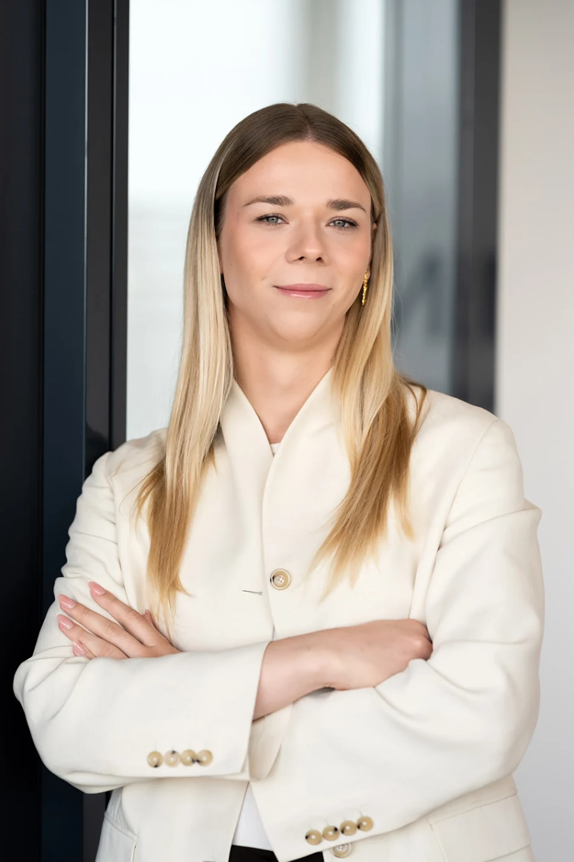 Frau mit langen blonden Haaren im weißen Blazer steht mit verschränkten Armen vor einer Glastür.