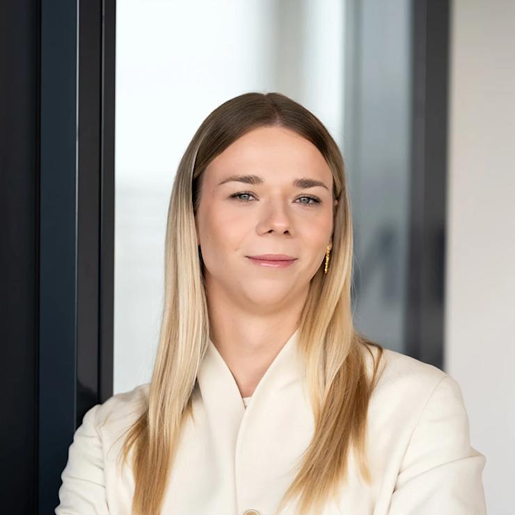 Frau mit langen blonden Haaren im weißen Blazer steht mit verschränkten Armen vor einer Glastür.