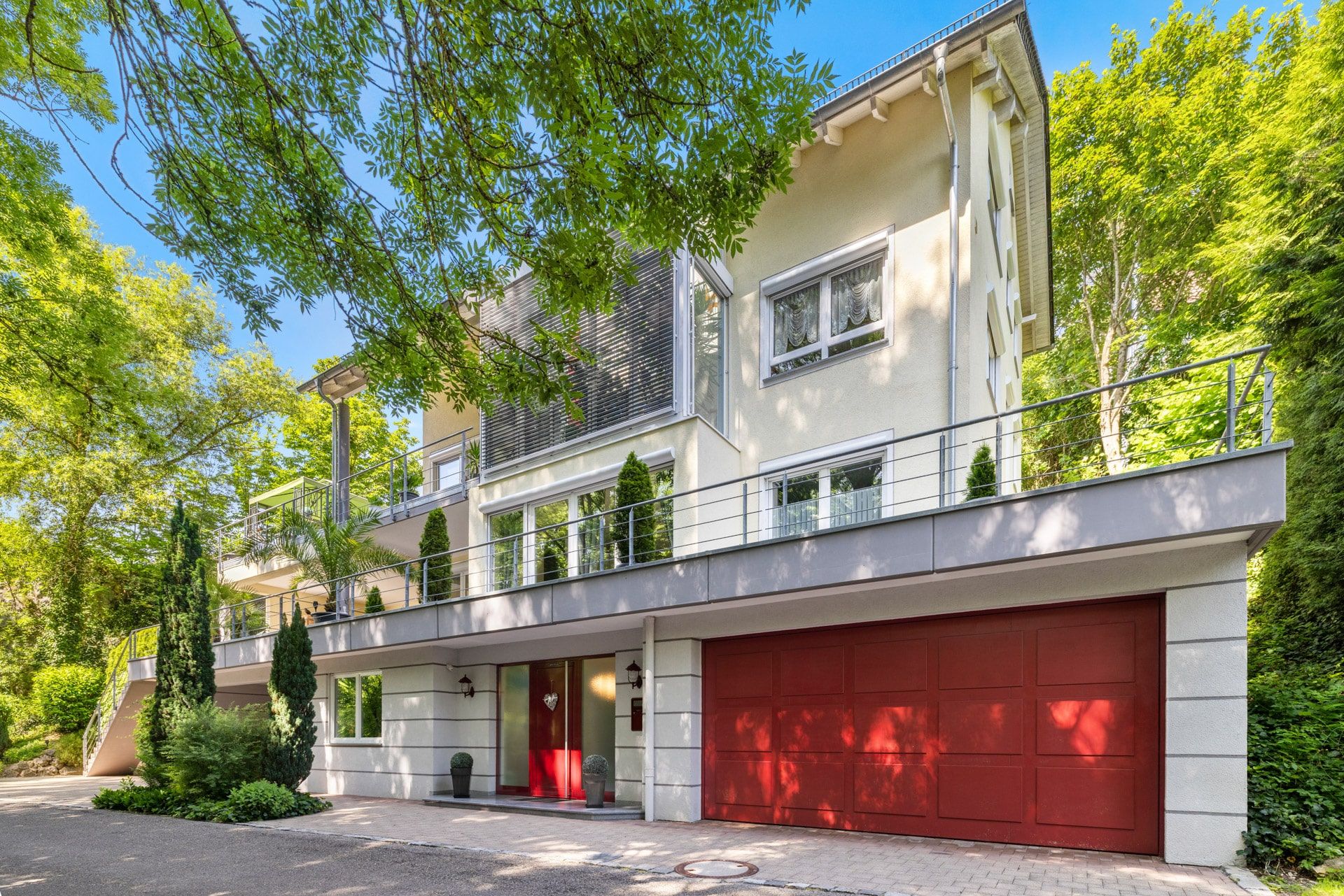 Modernes, dreistöckiges Wohnhaus mit roter Garage, Balkon und viel Grün im Vordergrund.
