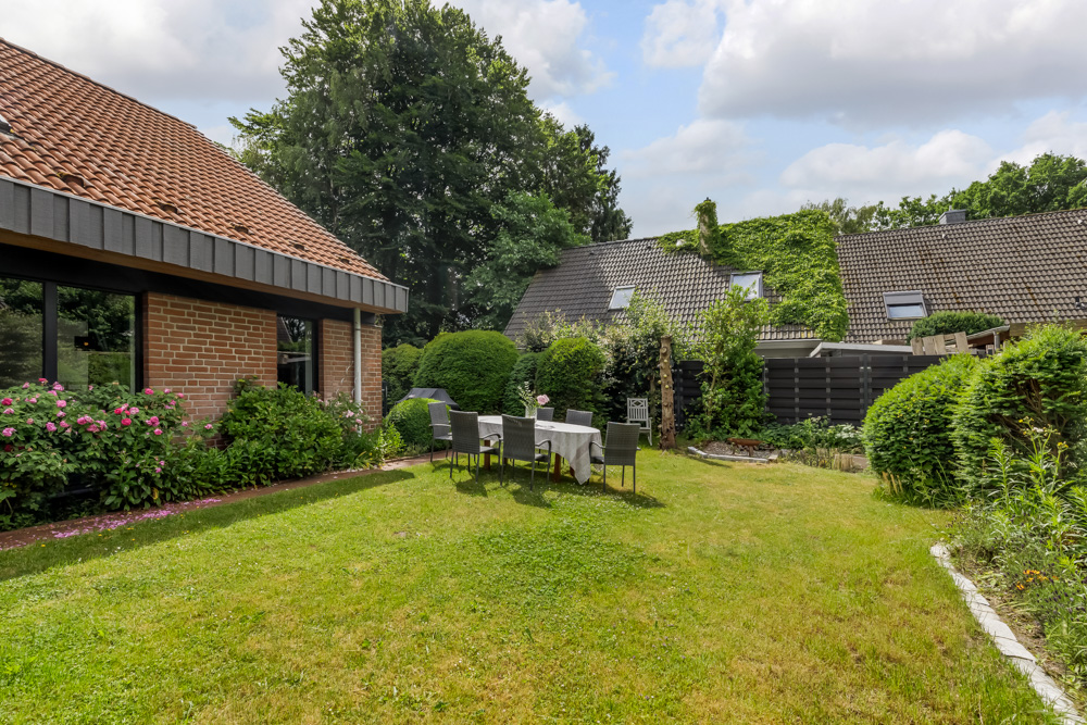 Garten mit Rasenfläche, Gartentisch und Stühlen neben einem Haus mit Blumen und Büschen.