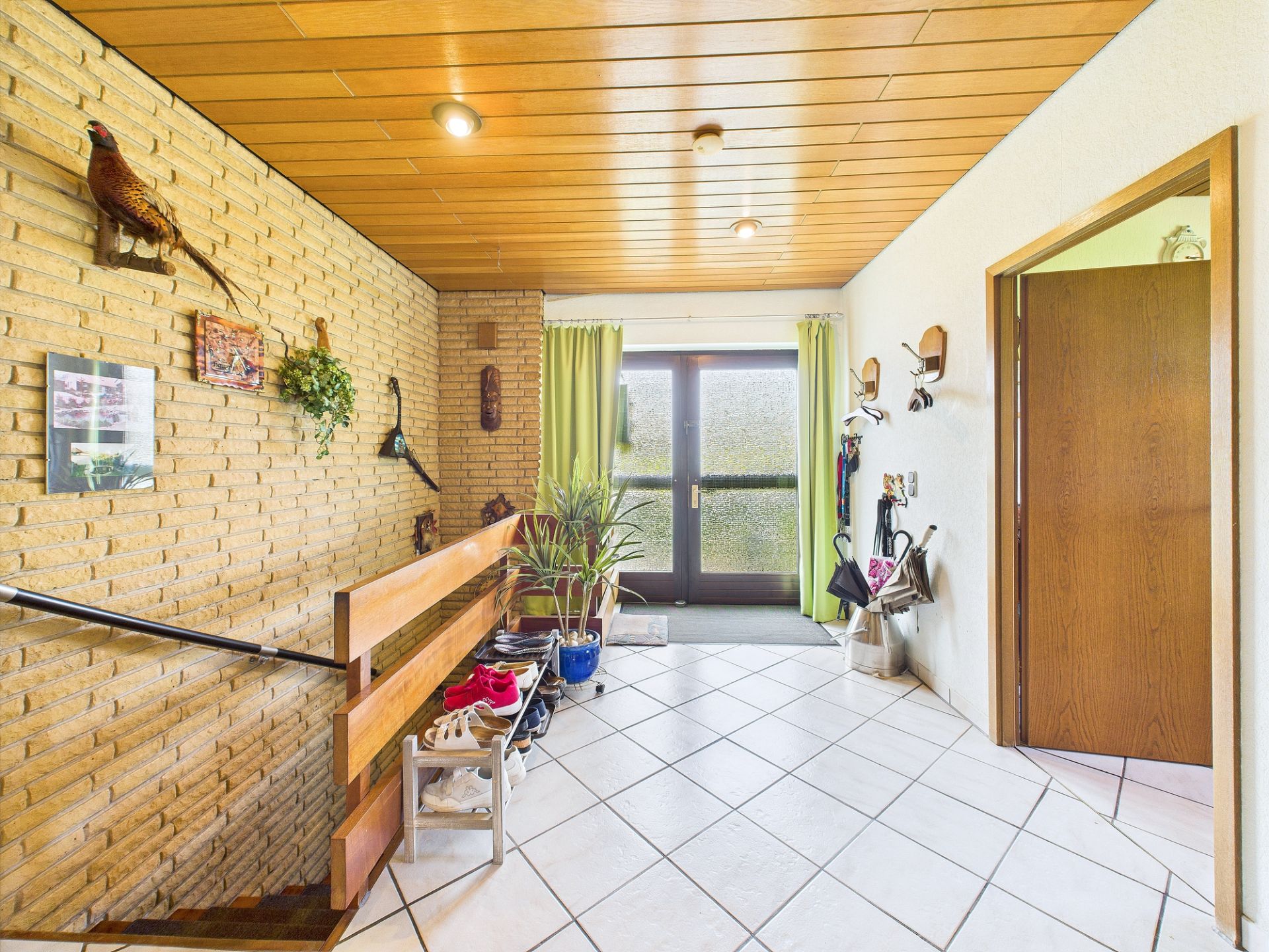 Heller Flur mit Fliesenboden, Holzwanddecke, Pflanzen und Schuhregal vor einer Glastür.