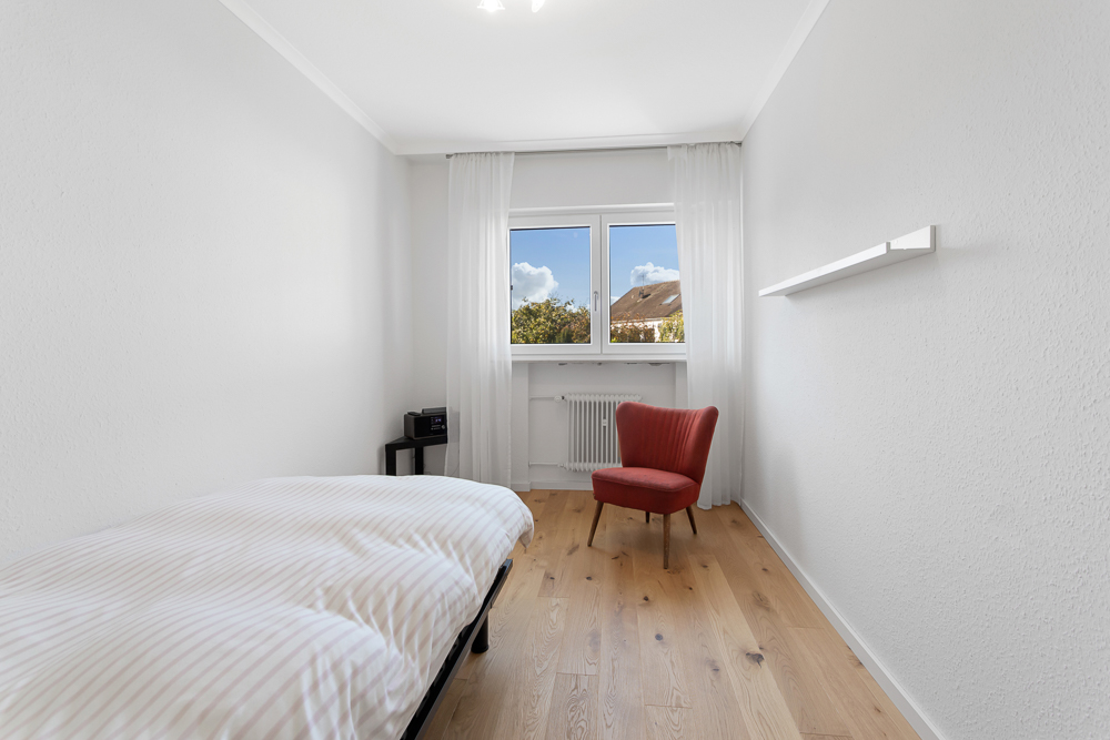 Helles Schlafzimmer mit Einzelbett, rotem Sessel und Fenster, Holzboden und weißen Wänden.