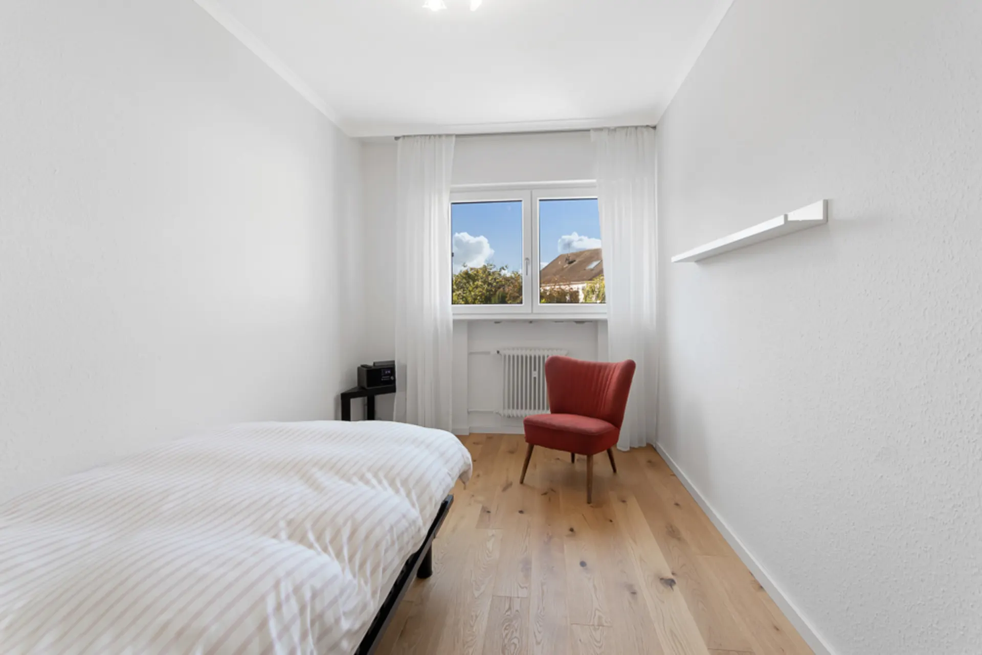 Helles Schlafzimmer mit Einzelbett, rotem Sessel und Fenster, Holzboden und weißen Wänden.