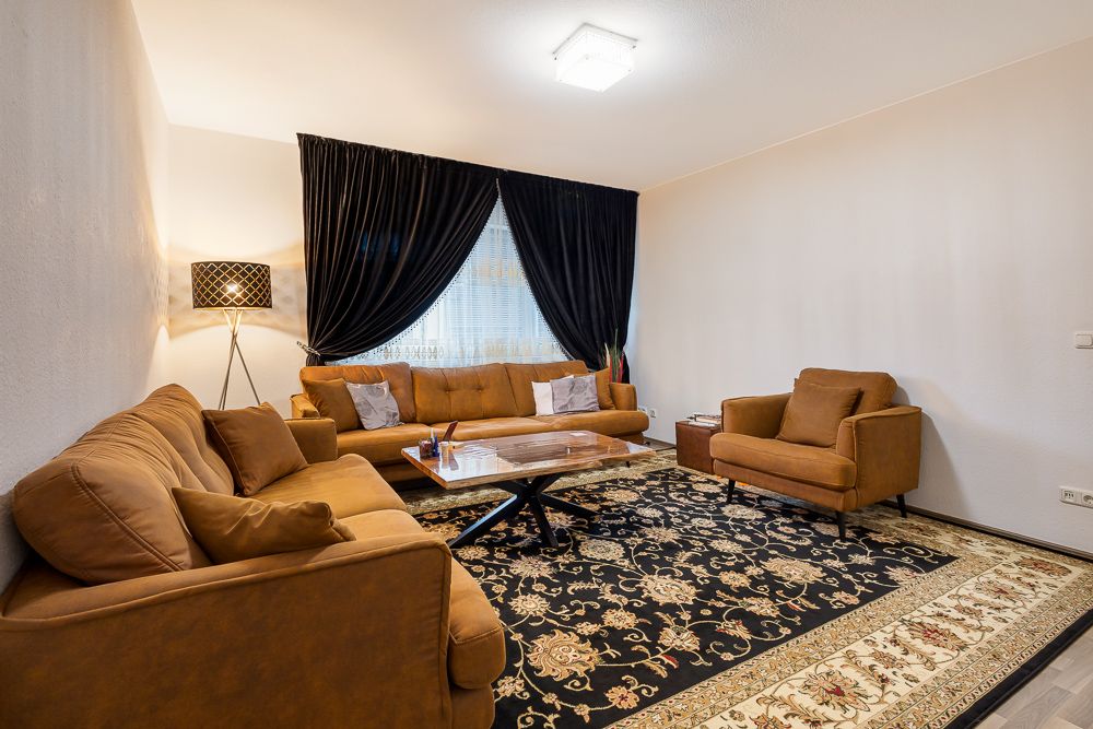 Wohnzimmer mit braunen Sofas, Couchtisch, Stehlampe und gemustertem Teppich vor dunklen Vorhängen.