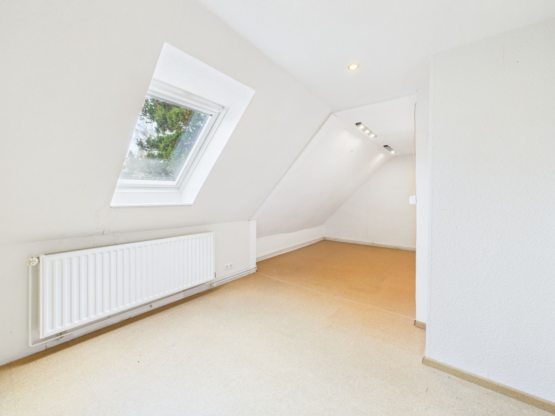 Helles Zimmer mit Dachschräge, großem Fenster und Heizung an der Wand.