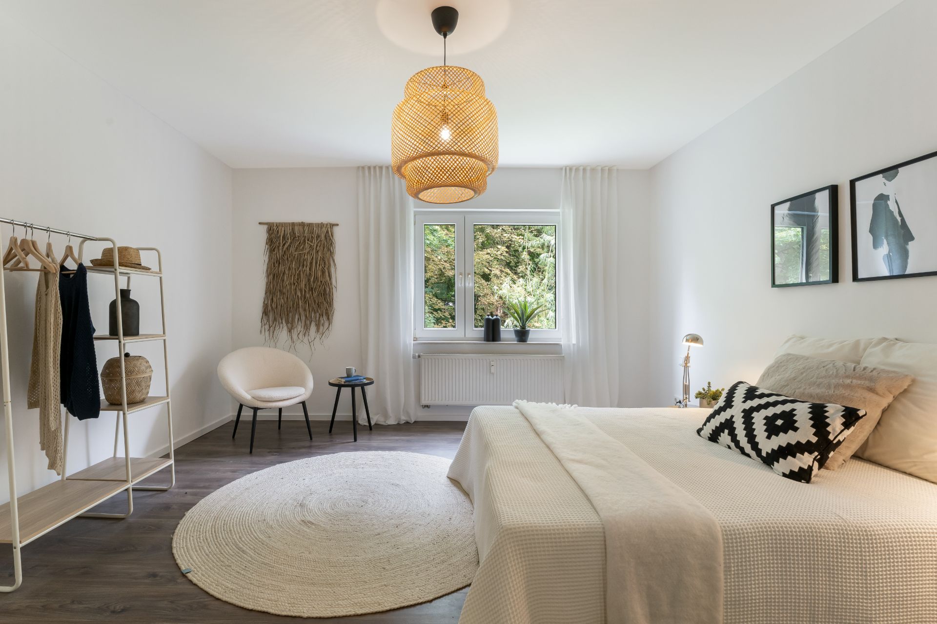 Helles Schlafzimmer mit Doppelbett, rundem Teppich, Fenster, offener Kleiderstange und Sessel.