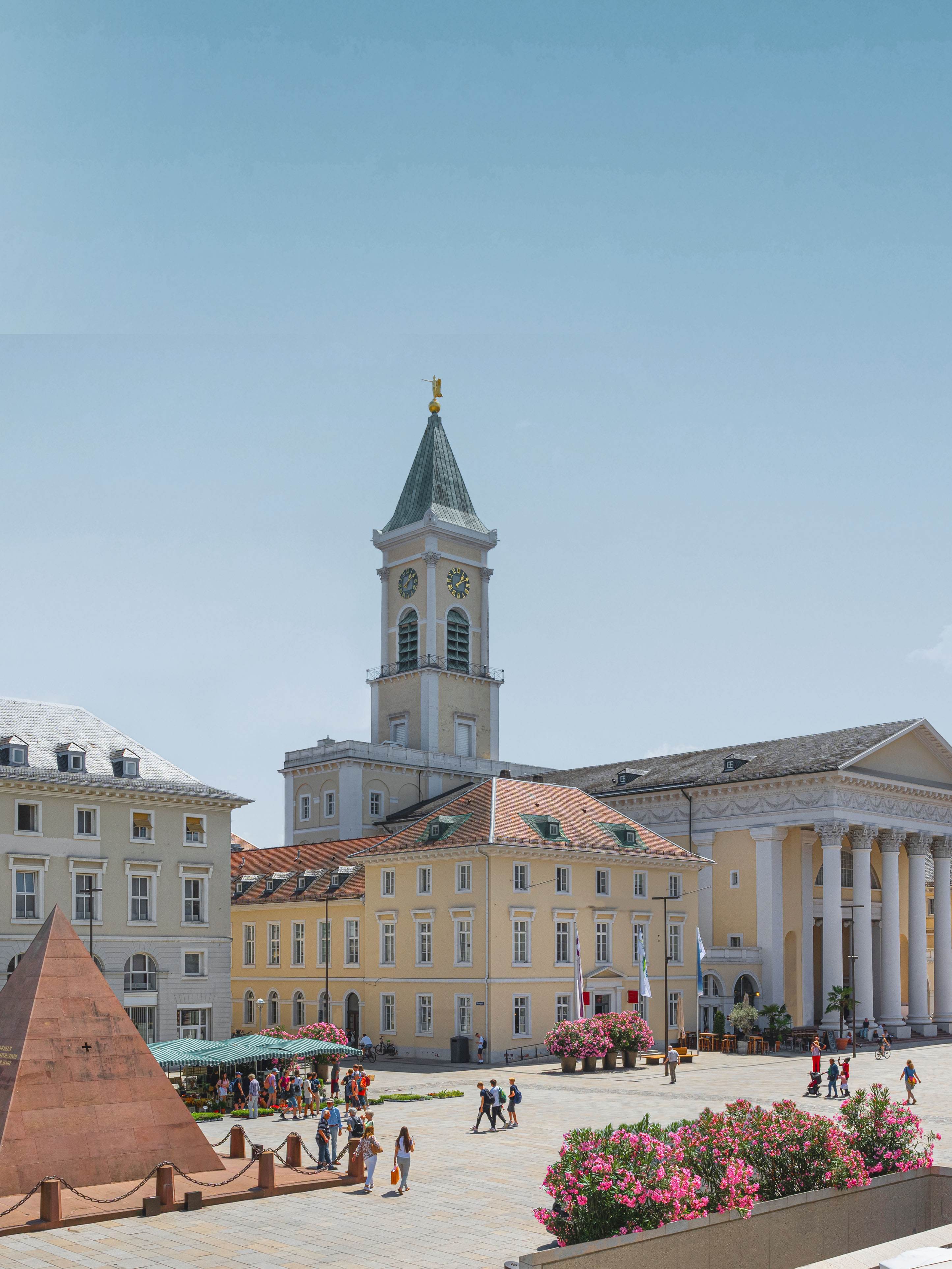 Stadtplatz mit Blumen, Pyramide, gelbem Gebäude und Uhrturm, Menschen spazieren.