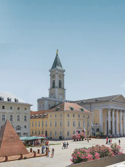 Stadtplatz mit Blumen, Pyramide, gelbem Gebäude und Uhrturm, Menschen spazieren.