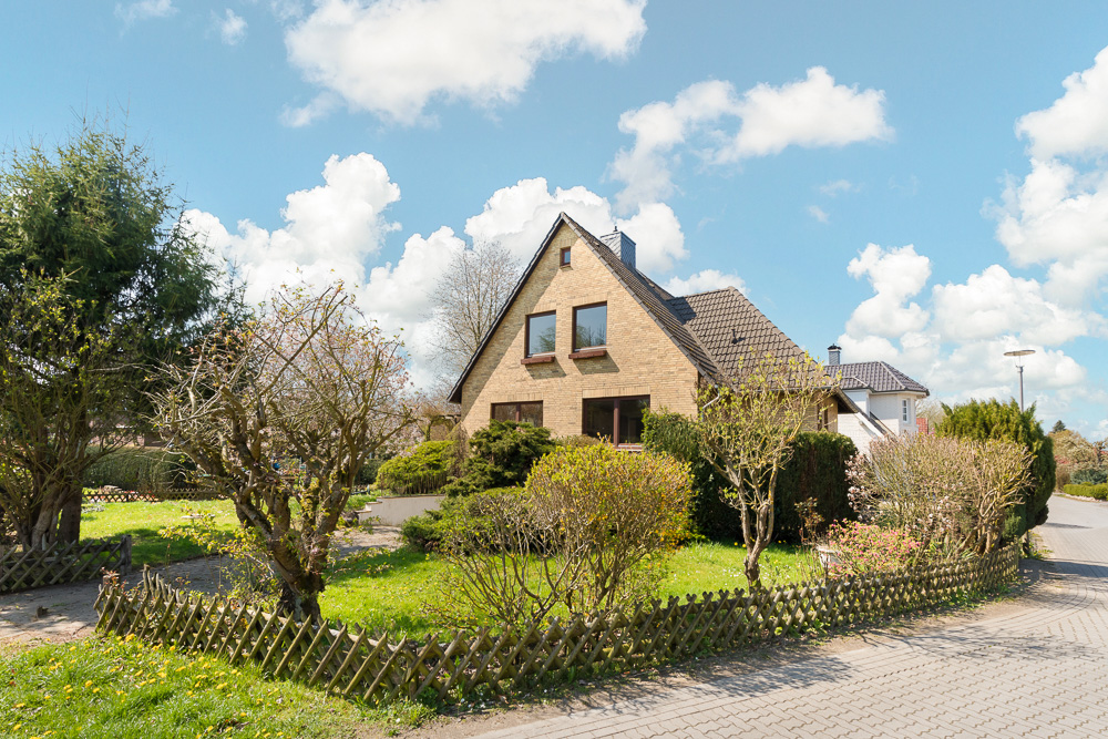 Einfamilienhaus mit Garten und Holzzaun an einer ruhigen Straße unter blauem Himmel.