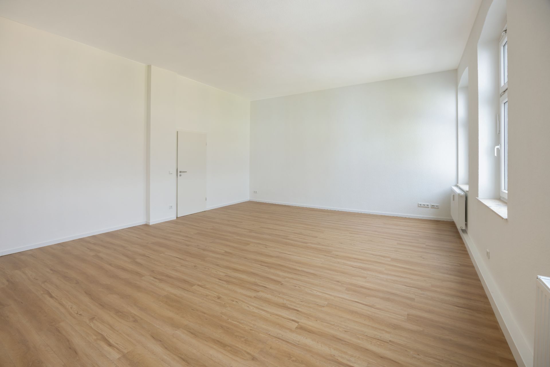 Leerer, heller Raum mit Holzboden, weißen Wänden und mehreren Fenstern.