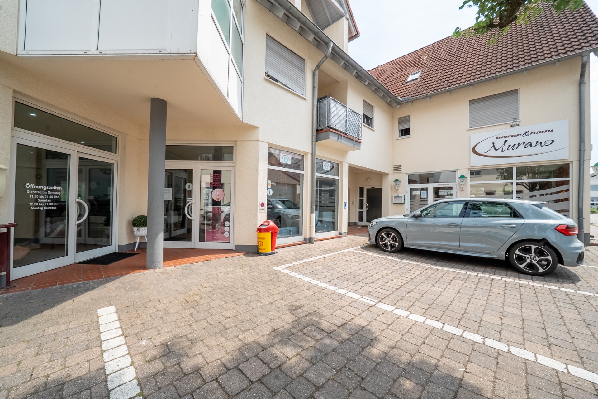 Außenansicht eines Wohnhauses mit Parkplatz, Auto und Ladeneingang.