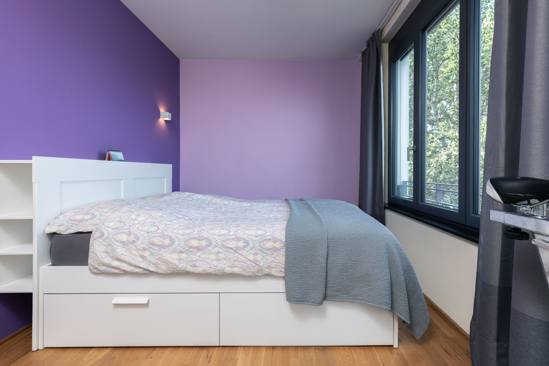 Schlafzimmer mit lila Wand, weißem Bett und großem Fenster mit grauen Vorhängen.