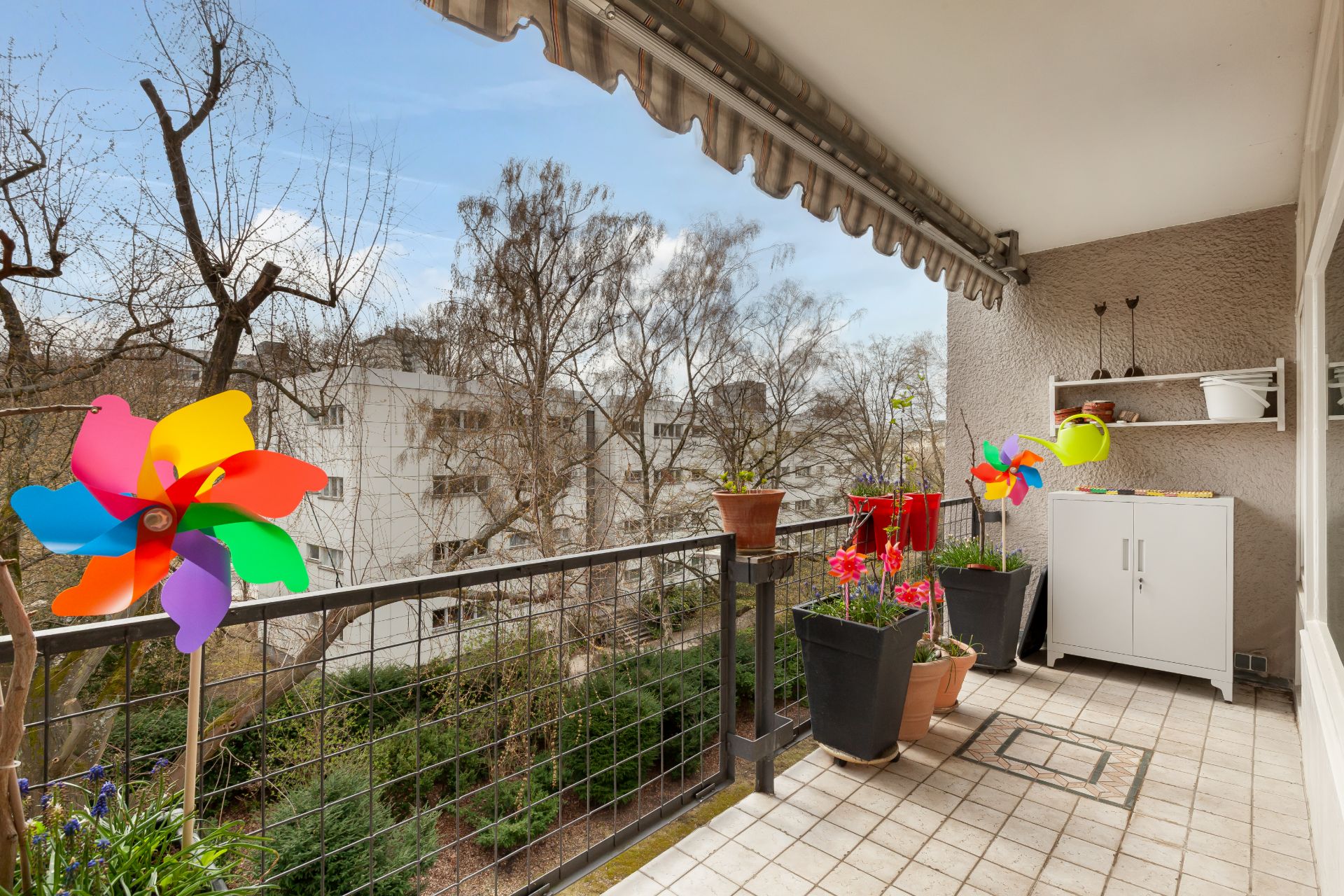 Balkon mit bunten Windrädern, Pflanzen und Schrank, darüber eine Markise.