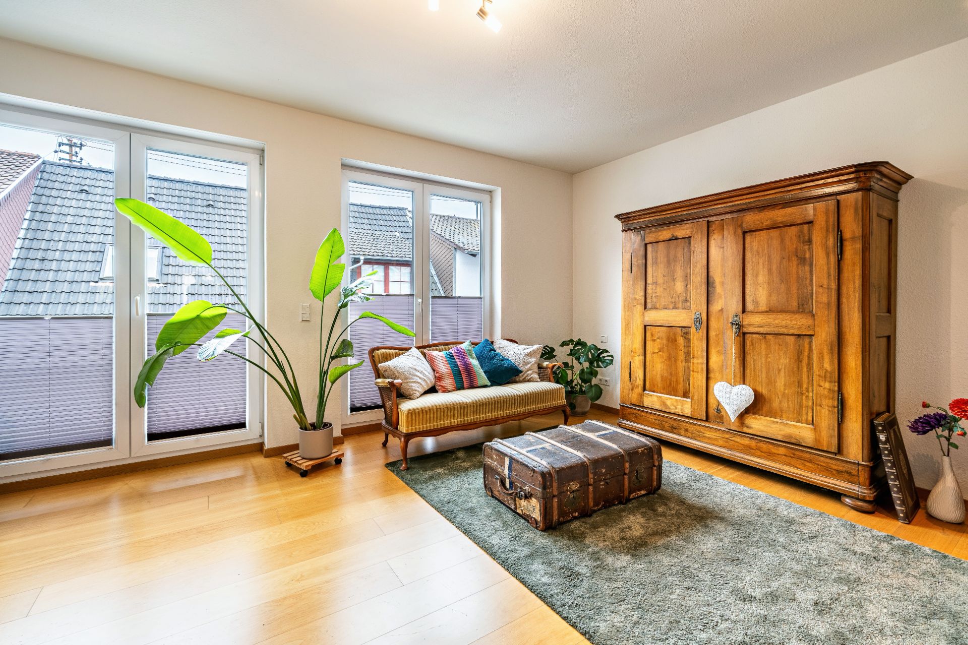 Wohnzimmer mit Holzboden, Sofa, großem Holzschrank, Pflanze und Truhe auf Teppich.