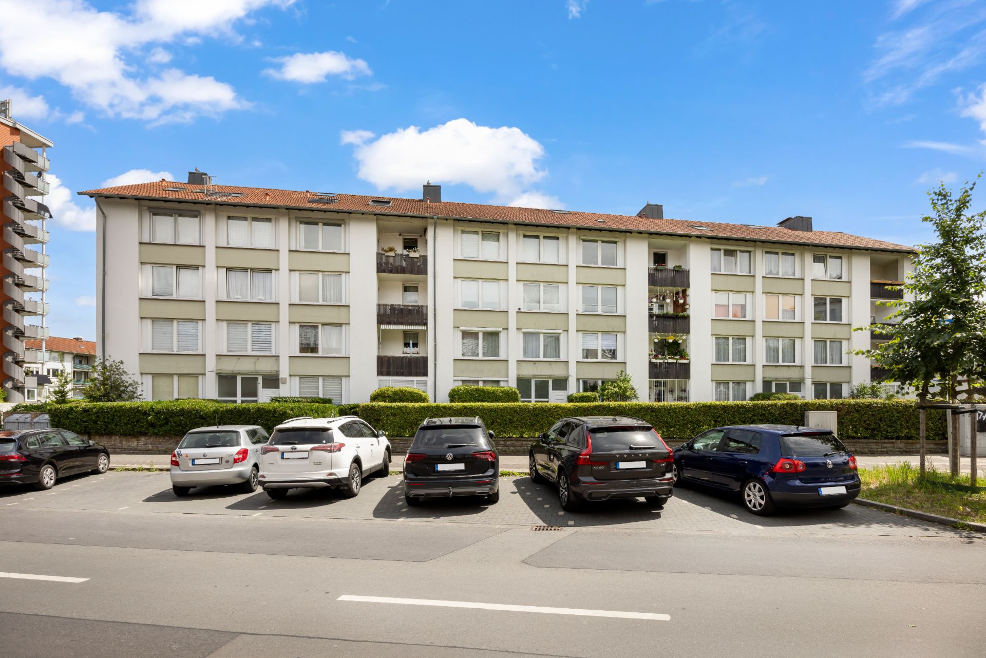 Mehrfamilienhaus mit vier Etagen und vorgelagertem Parkplatz mit geparkten Autos.