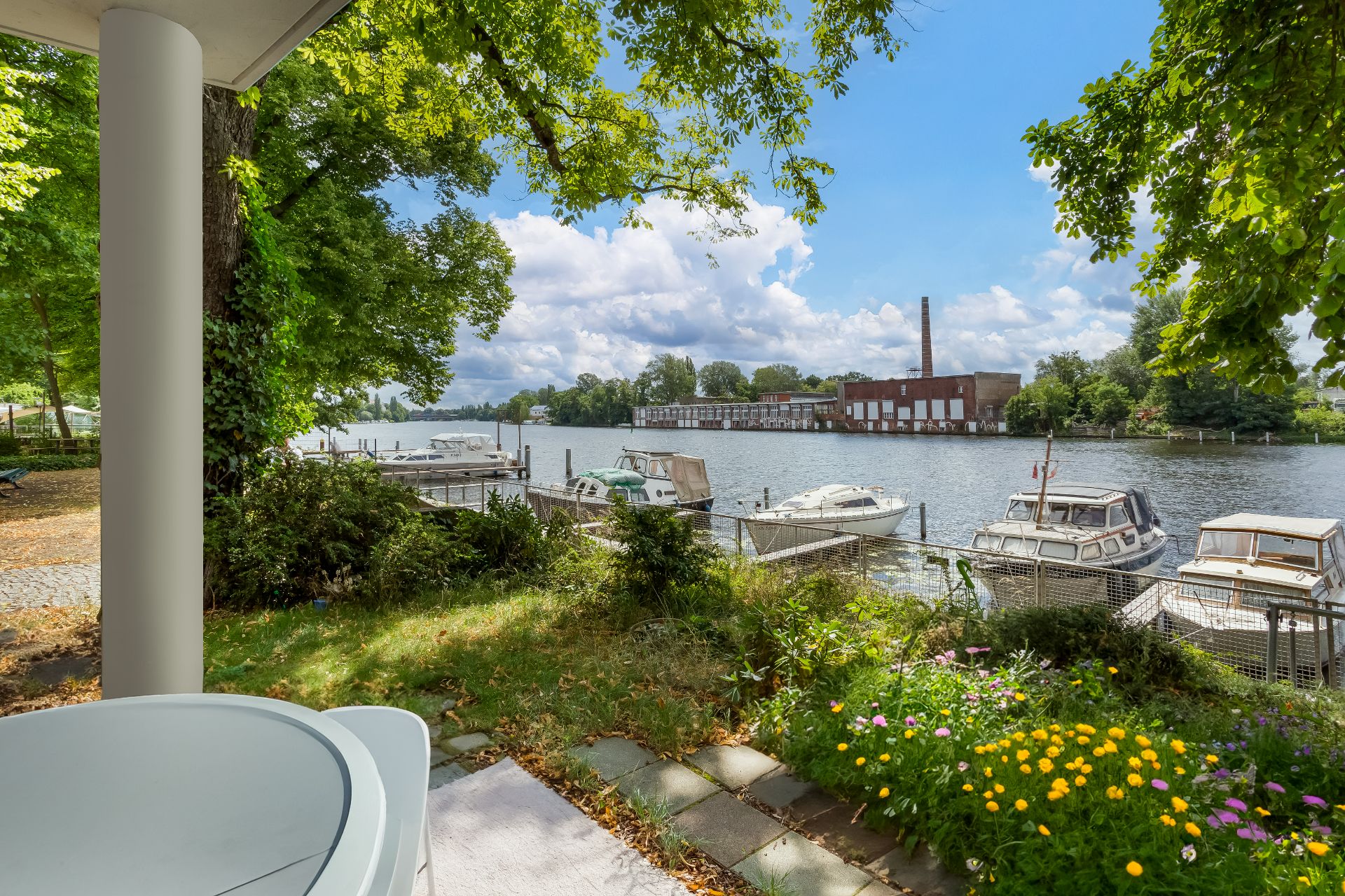 Blick von einer Terrasse auf einen Garten und einen Fluss mit Booten am Steg.