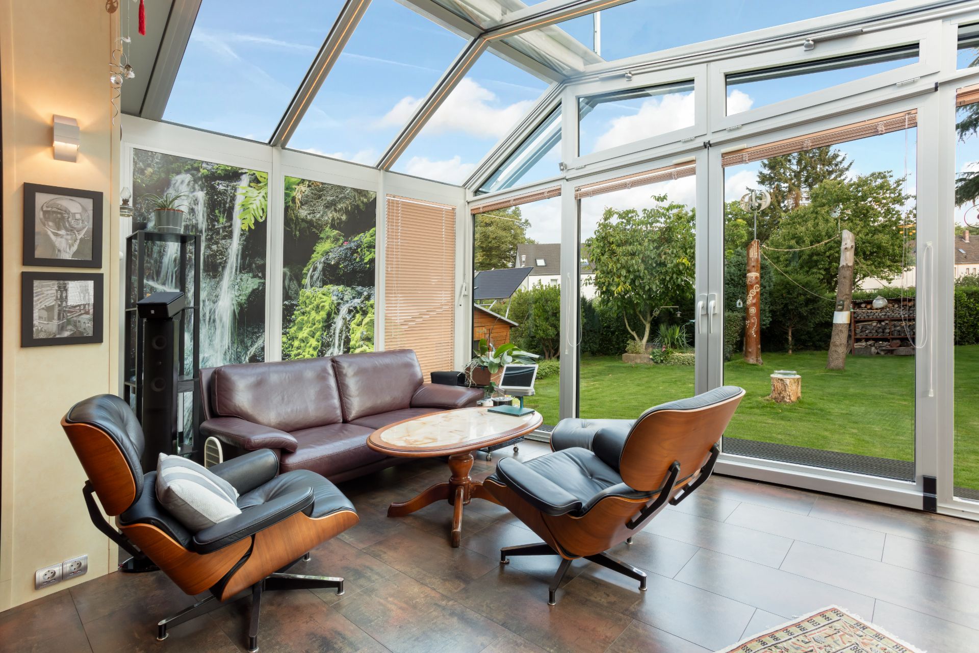 Heller Wintergarten mit Ledersofa, zwei Sesseln und Couchtisch, Blick in den grünen Garten.