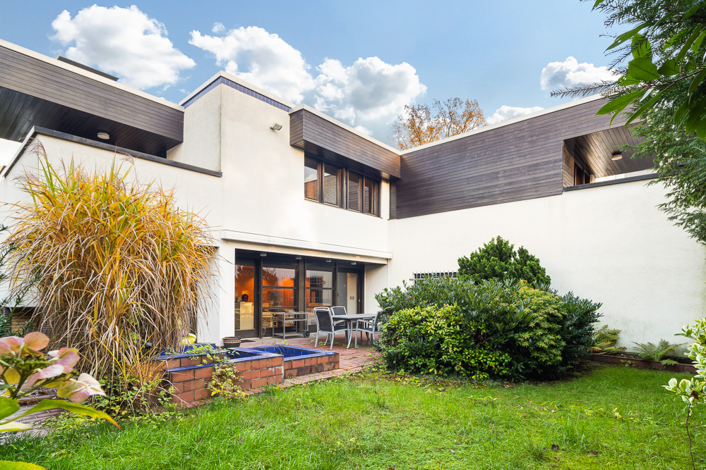 Moderner Garten mit Terrasse, Sitzbereich, Pflanzen und Blick auf ein weißes Wohnhaus.
