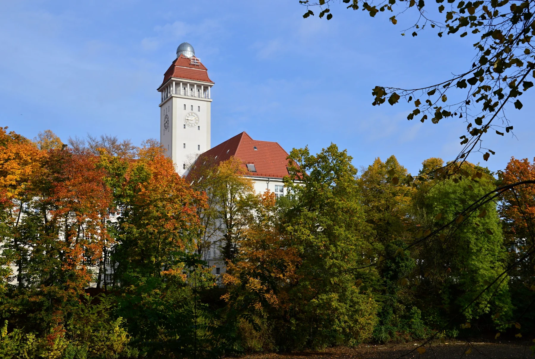 Weißes Gebäude mit Uhrturm hinter bunten Herbstbäumen unter blauem Himmel.