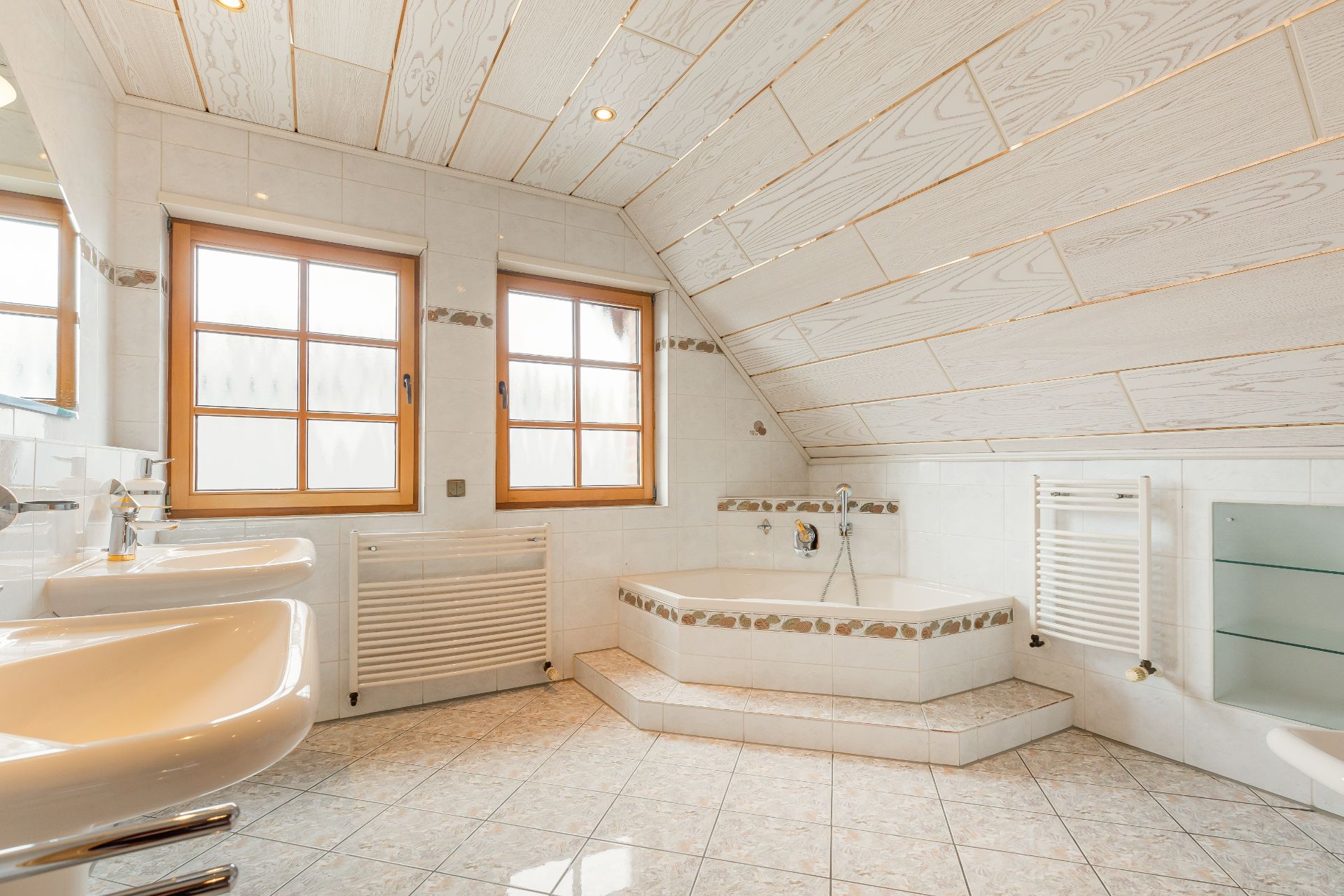 Helles Badezimmer mit zwei Fenstern, Eckbadewanne, zwei Waschbecken und schräger Holzdecke.