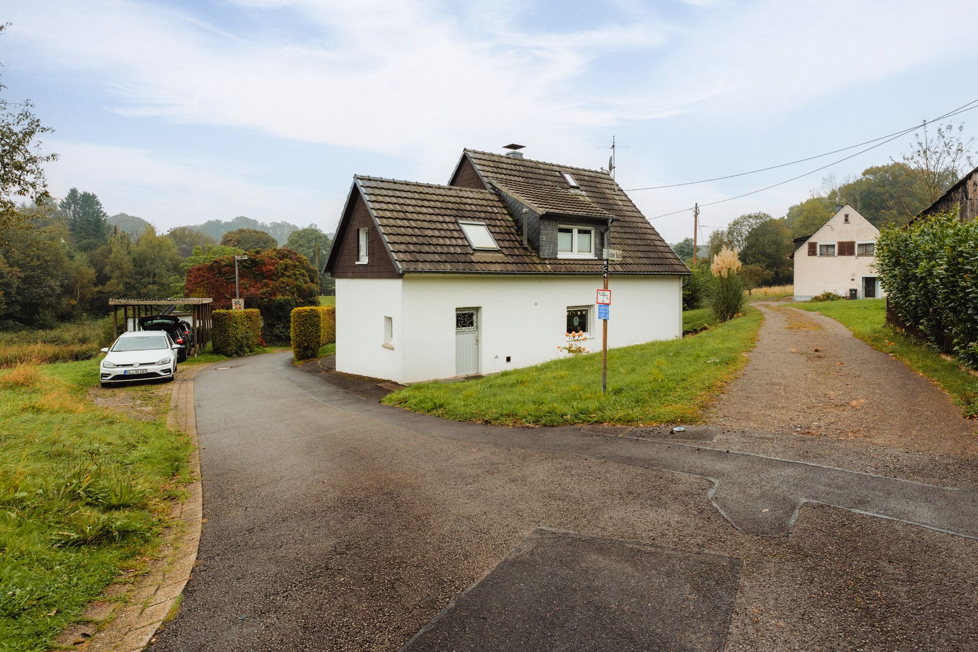Weißes Einfamilienhaus mit Garten an einer Weggabelung, Auto steht links unter Carport.