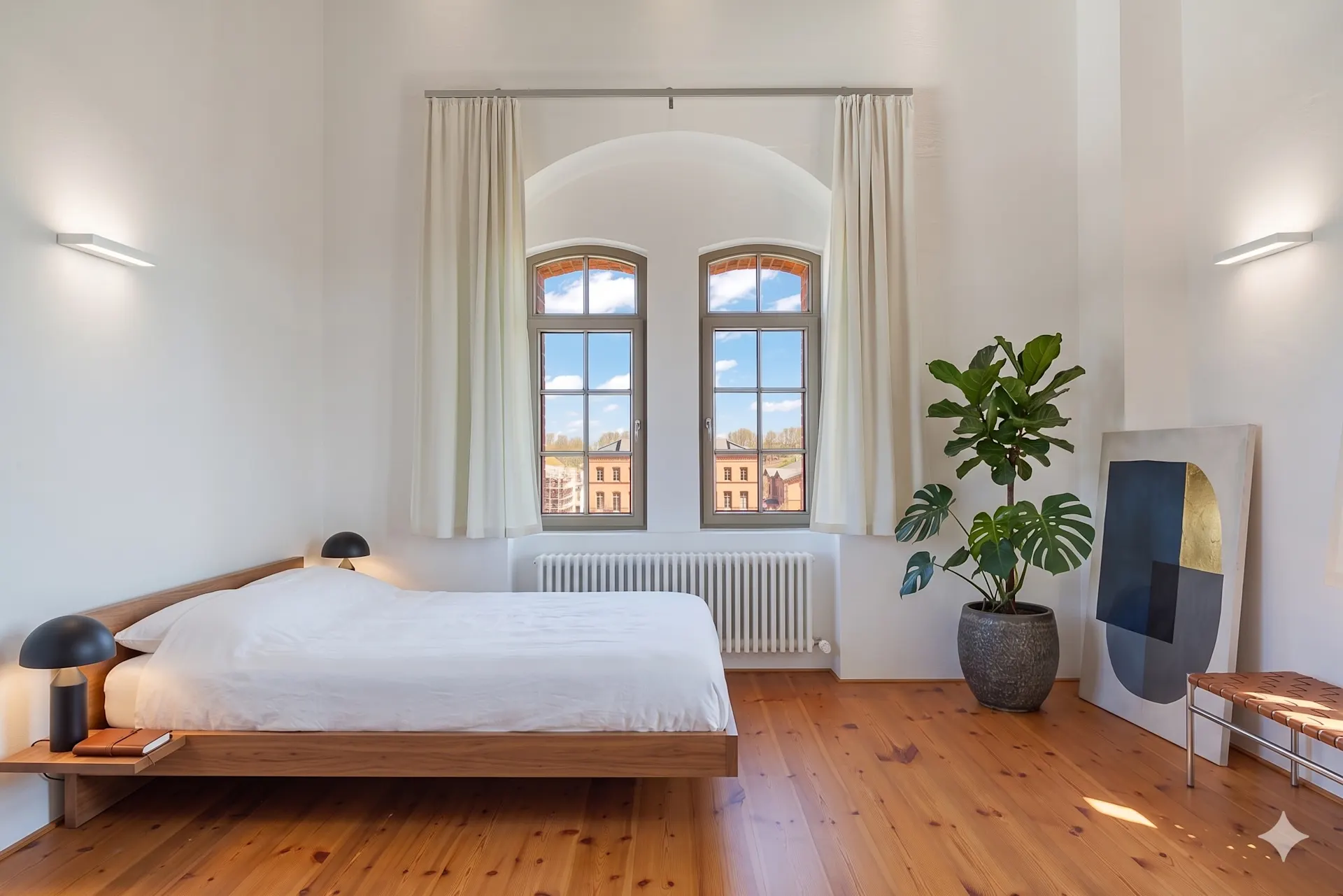 Helles Schlafzimmer mit Holzboden, Bett, zwei Lampen, großem Fenster, Pflanze und moderner Kunst.