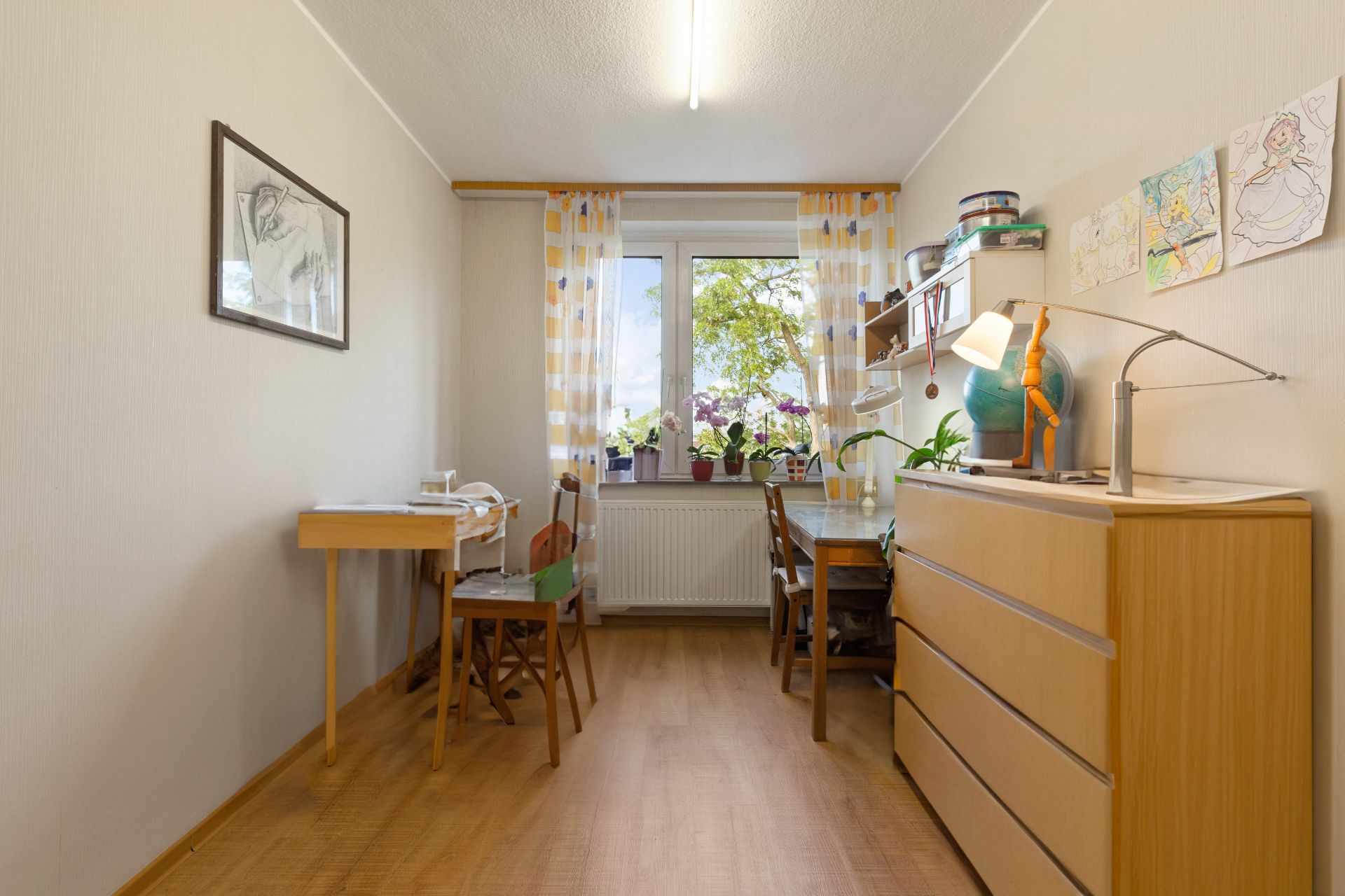 Helles Arbeitszimmer mit Schreibtisch, Kommode und Fenster mit Blick auf grüne Bäume.