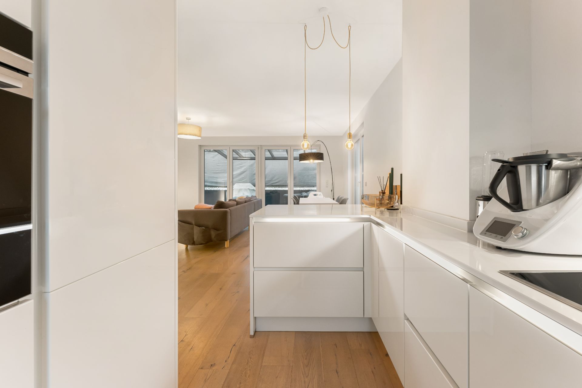 Moderne weiße Küche mit Holzboden und Blick auf Wohnzimmer mit großen Fenstern.