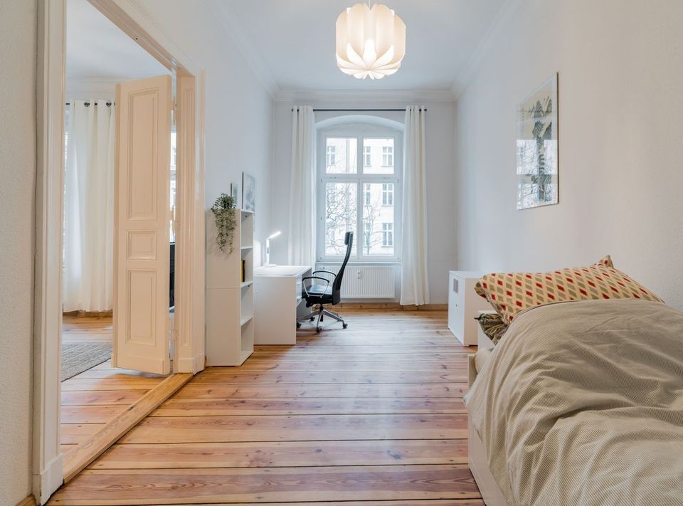 Helles Zimmer mit Einzelbett, Schreibtisch und Holzfußboden, großes Fenster mit weißen Vorhängen.