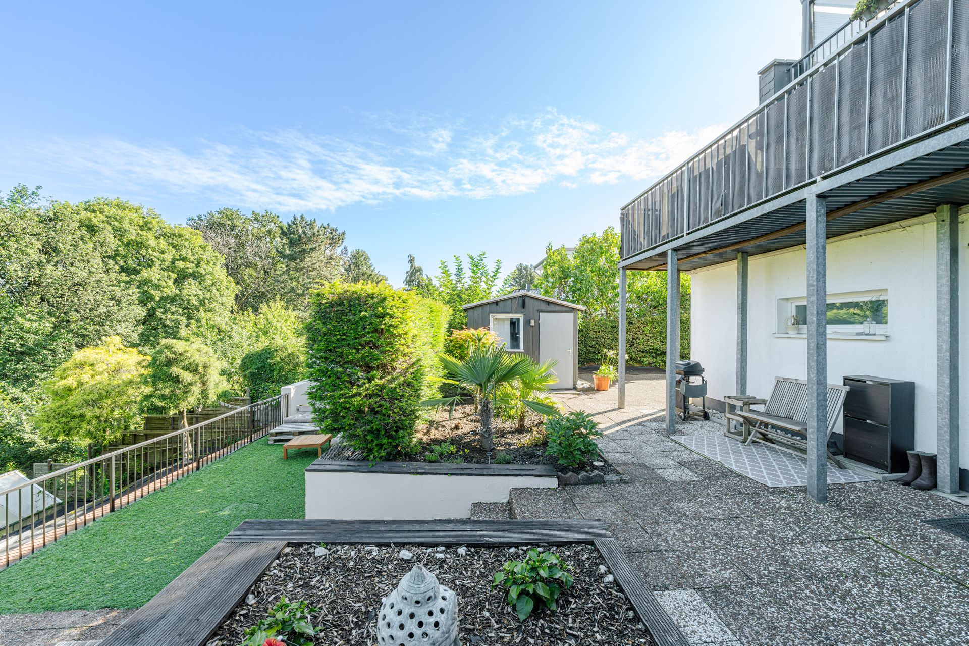 Gepflegter Garten mit Terrasse, Sitzbank, Grill und Gartenhaus; umgeben von vielen Bäumen.