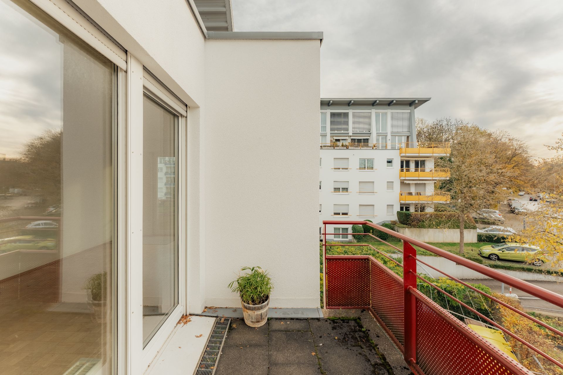 Balkon mit rotem Geländer, Pflanze im Topf und Blick auf Wohngebäude im Hintergrund.