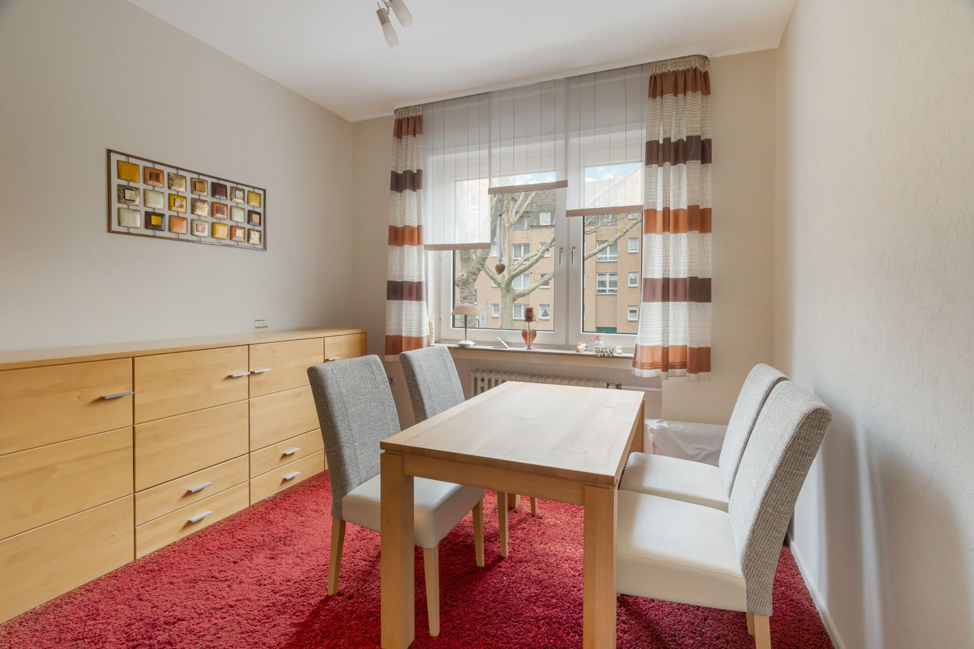 Esszimmer mit Holztisch, vier Stühlen, rotem Teppich und Kommode vor einem Fenster mit bunten Vorhängen.