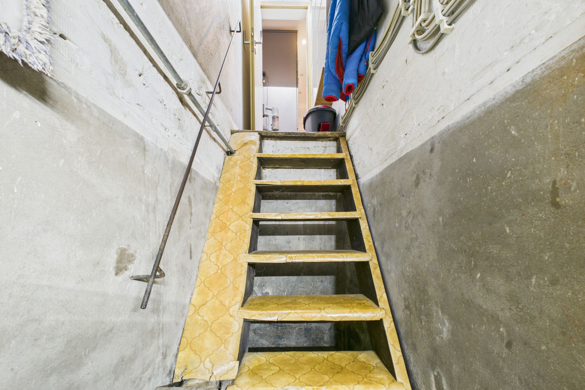 Schmale Kellertreppe mit gelbem Teppich, Handlauf und sichtbaren Wänden.