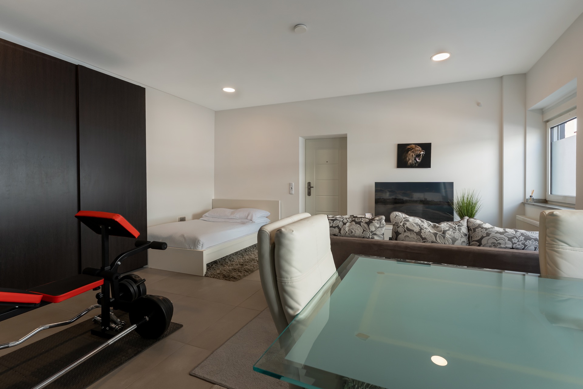 Helles Wohnstudio mit Bett, Sofa, Fernseher, Glastisch und Fitnessgerät.