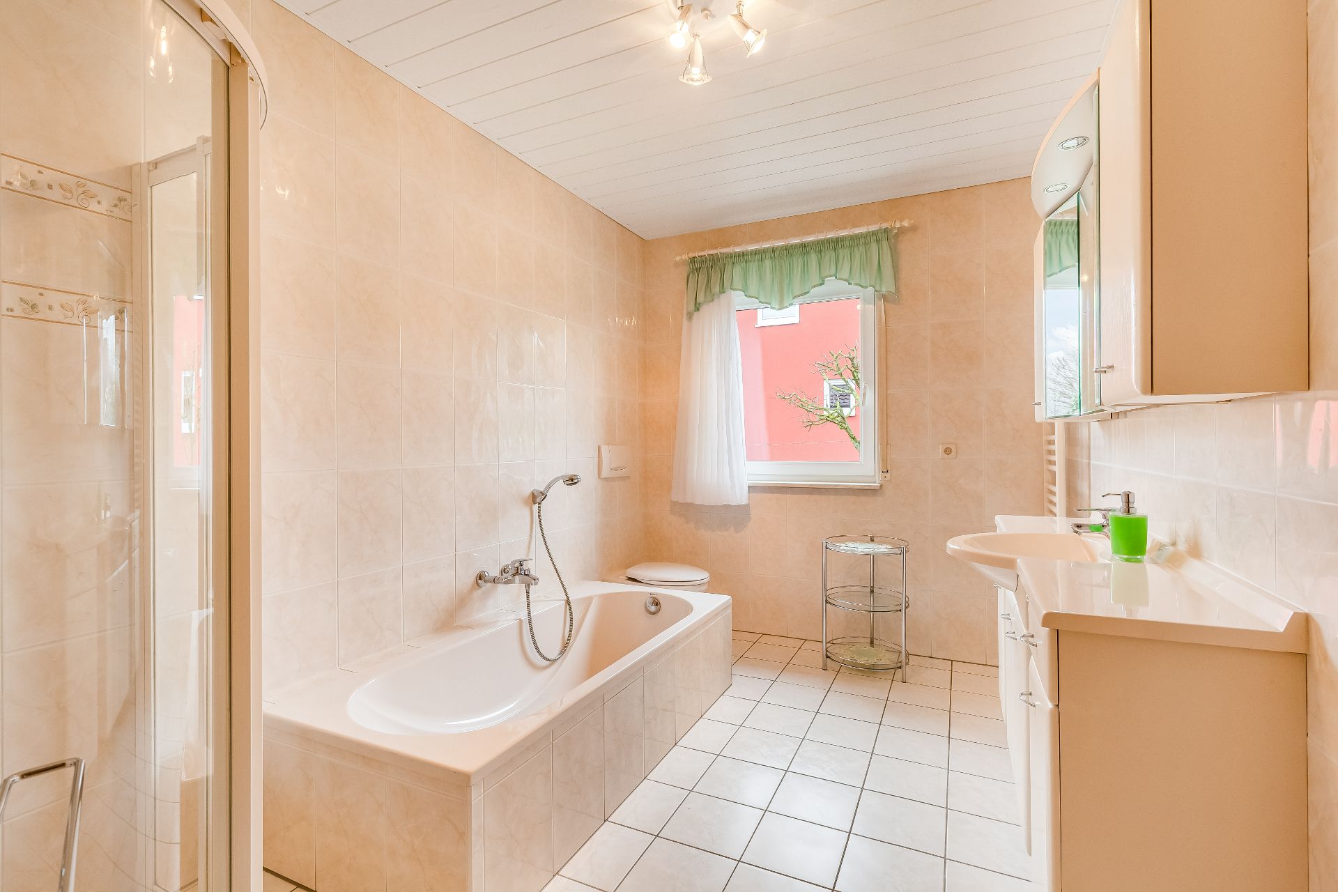 Helles Badezimmer mit Badewanne, Dusche, Waschbecken, WC und Fenster mit Vorhang.
