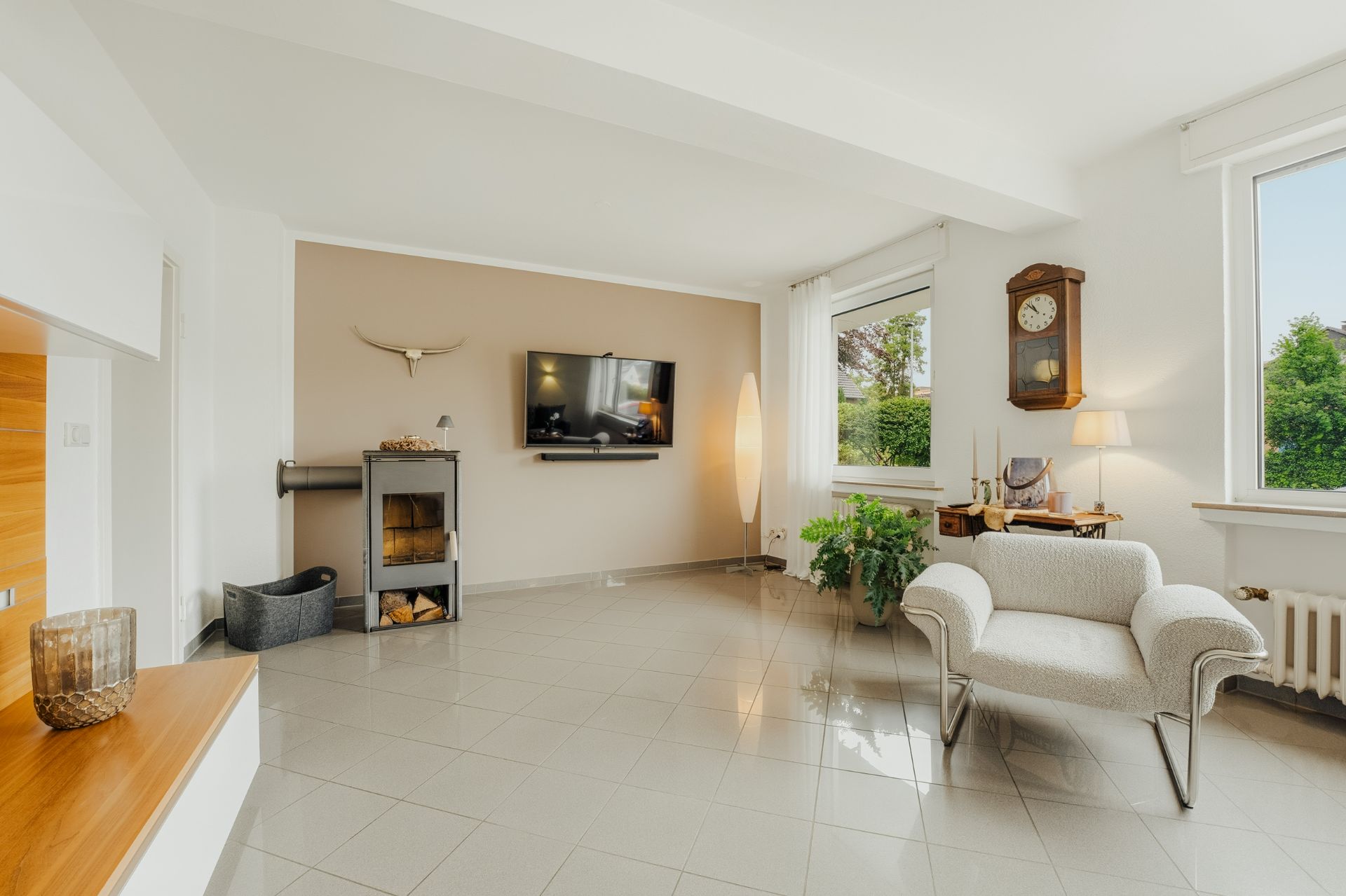 Helles Wohnzimmer mit Sessel, Fernseher, Kamin, großen Fenstern und einer Standuhr an der Wand.