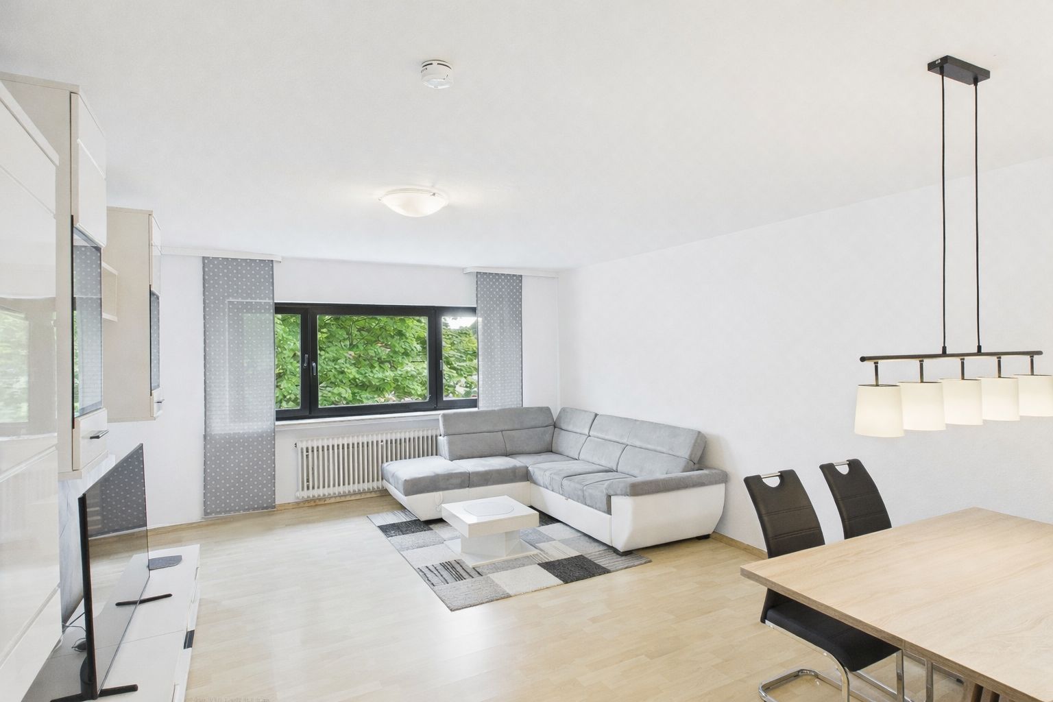 Helles Wohnzimmer mit Ecksofa, Teppich, Esstisch und großen Fenstern mit Blick ins Grüne.