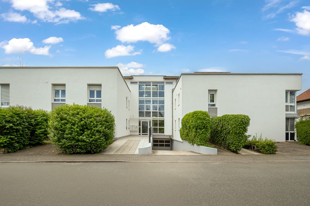 Modernes weißes Wohngebäude mit großen Fenstern und gepflegten Büschen am Eingang.