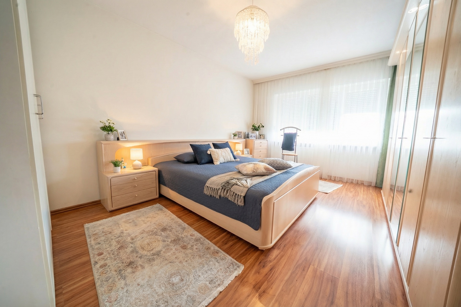 Helles Schlafzimmer mit Doppelbett, Holzboden, großem Schrank und Fenster mit Gardinen.