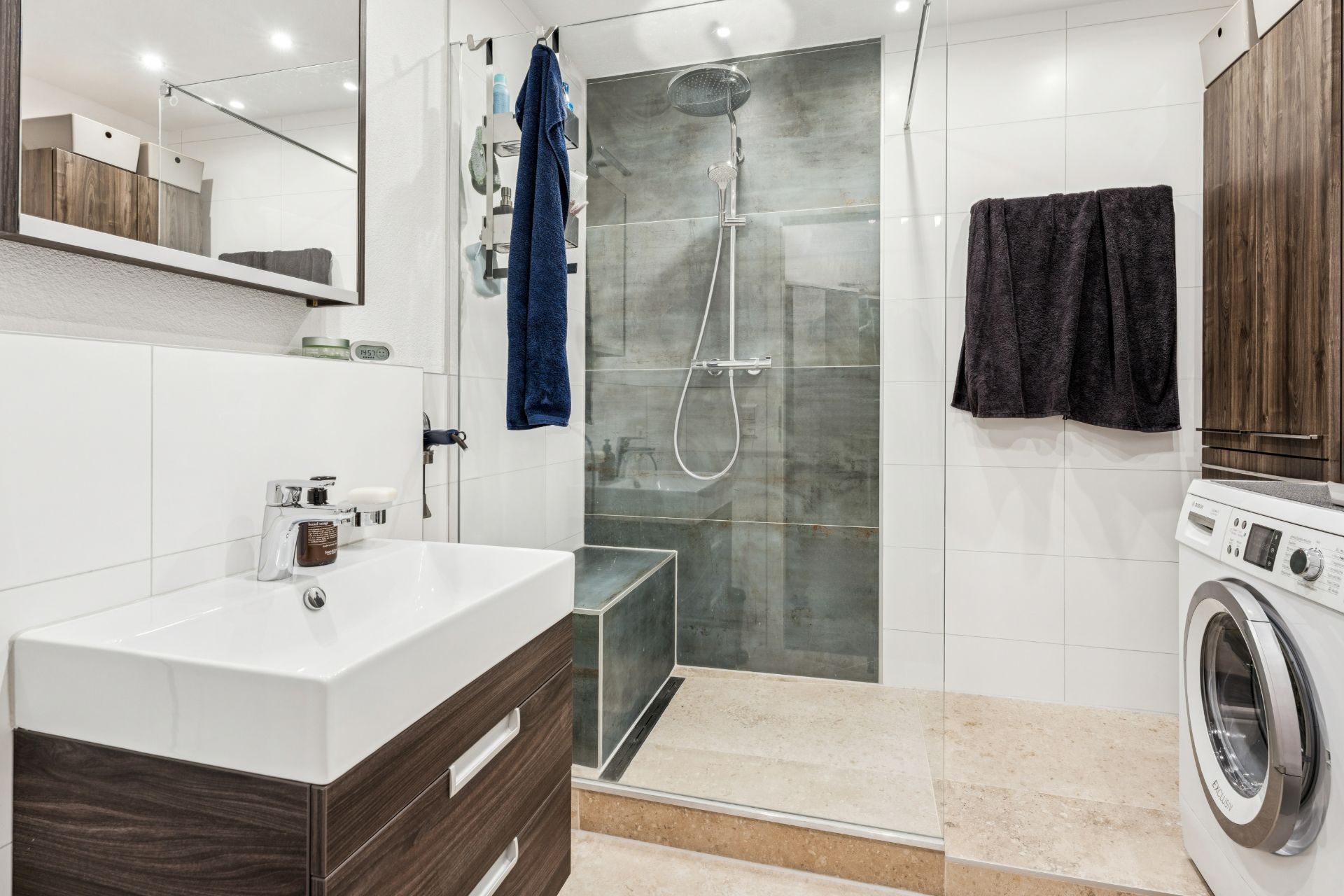 Modernes Badezimmer mit Waschbecken, Dusche, Waschmaschine und dunklen Handtüchern an der Wand.