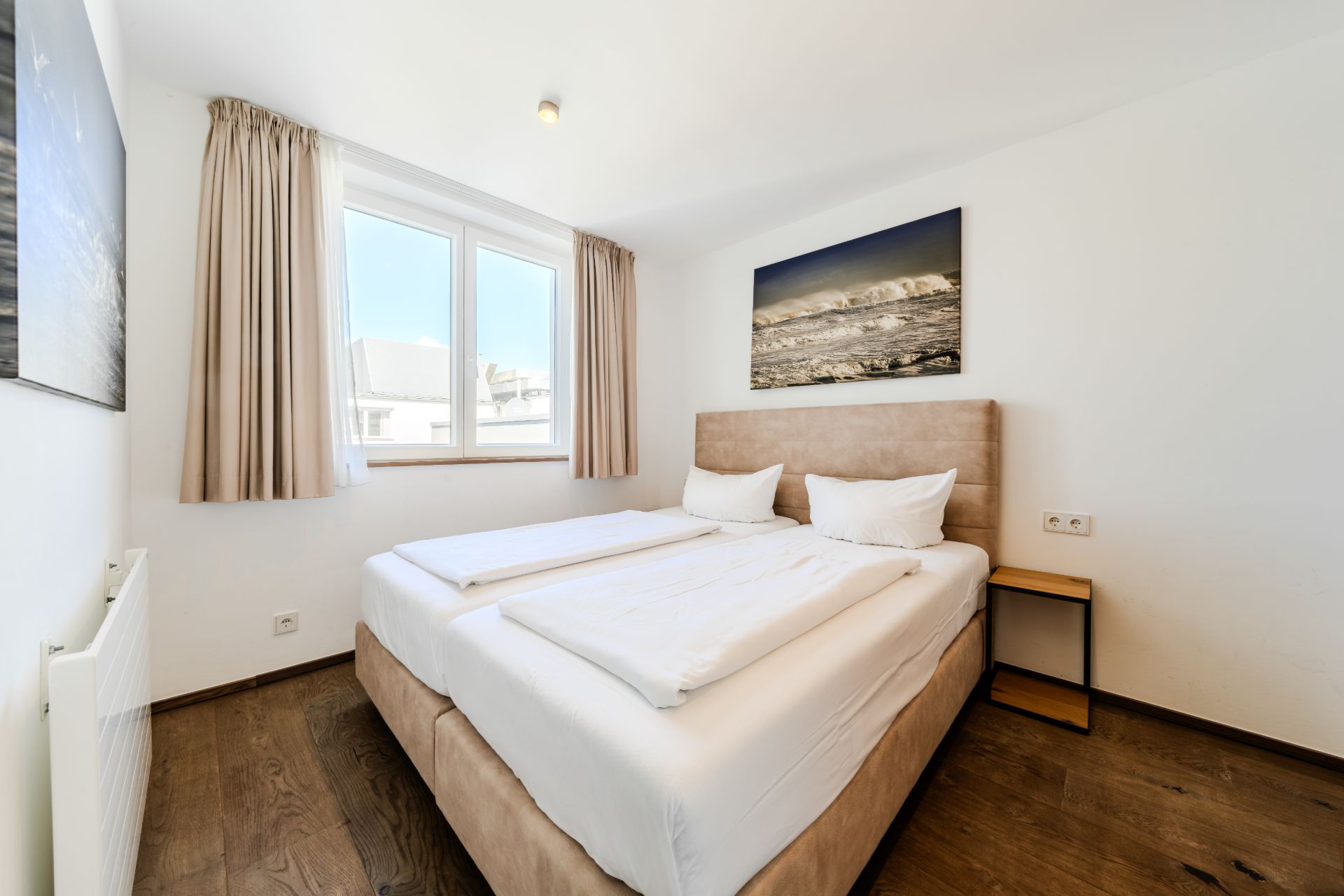 Helles Schlafzimmer mit Doppelbett, Nachttisch, großem Fenster und Bild an der Wand.