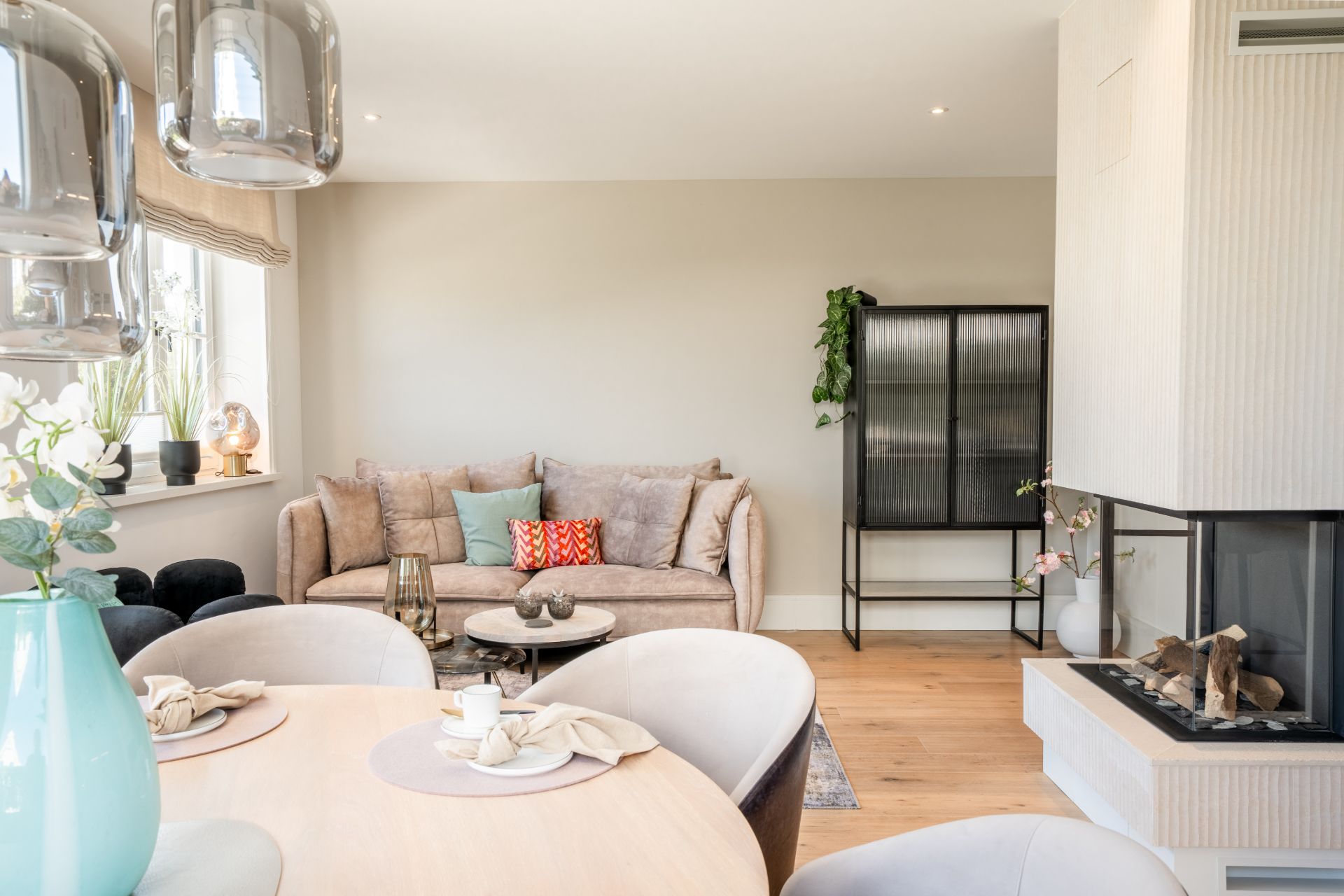 Helles Wohnzimmer mit Sofa, Kamin, Esstisch und großen Fenstern mit Pflanzen.