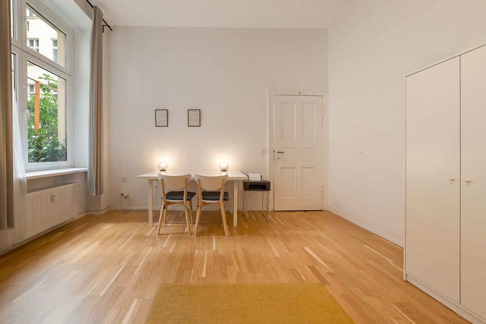 Helles Zimmer mit Holzboden, Tisch und zwei Stühlen, großem Fenster und weißem Kleiderschrank.