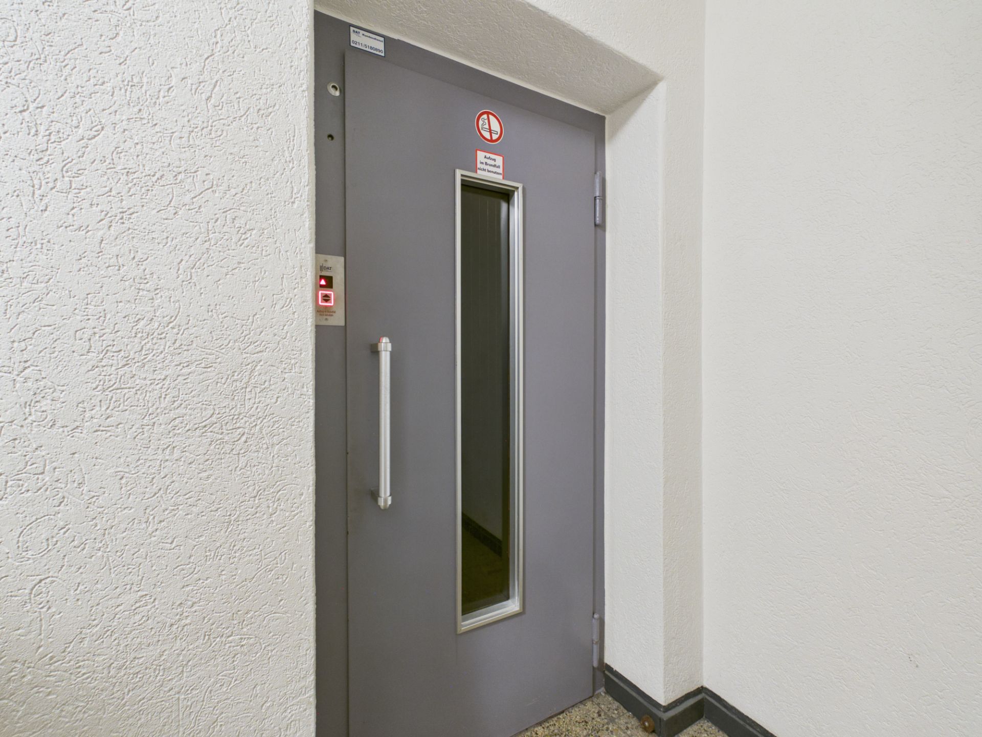 Graue Aufzugtür mit schmalem Fenster und silbernem Griff im Flur eines Wohngebäudes.